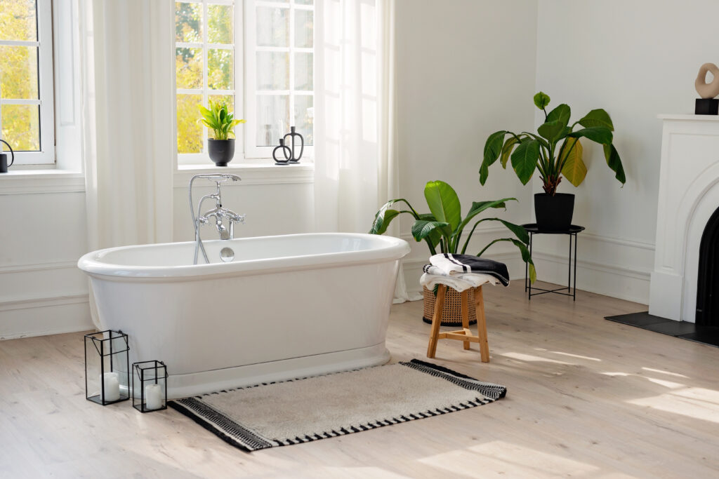 Stylish modern bathroom interior. Horizontal view of an empty free-standing bathtub on a wooden floor in a bright room against the backdrop of a large window and houseplants. Soft selective focus.